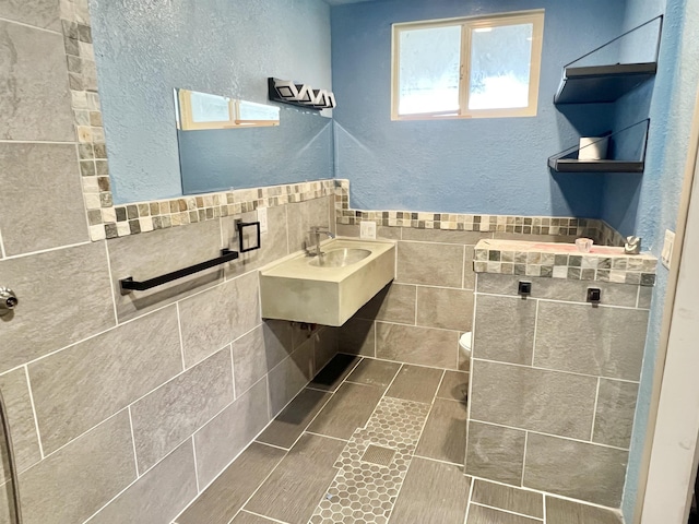 bathroom featuring toilet, wainscoting, a textured wall, tile walls, and a sink