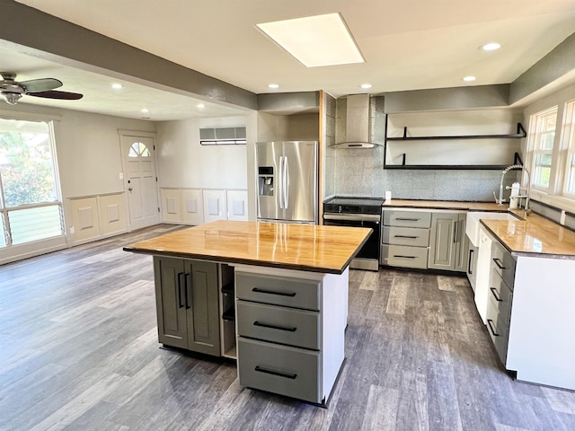 kitchen with gray cabinetry, open shelves, stainless steel appliances, butcher block counters, and wall chimney range hood