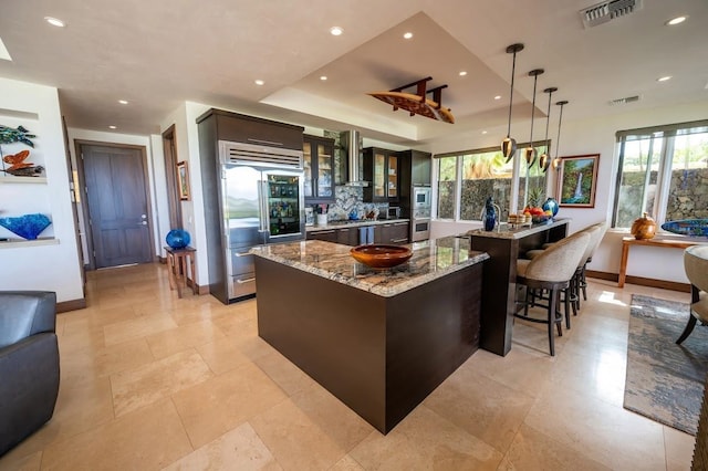 kitchen with appliances with stainless steel finishes, a center island, wall chimney range hood, hanging light fixtures, and light stone counters