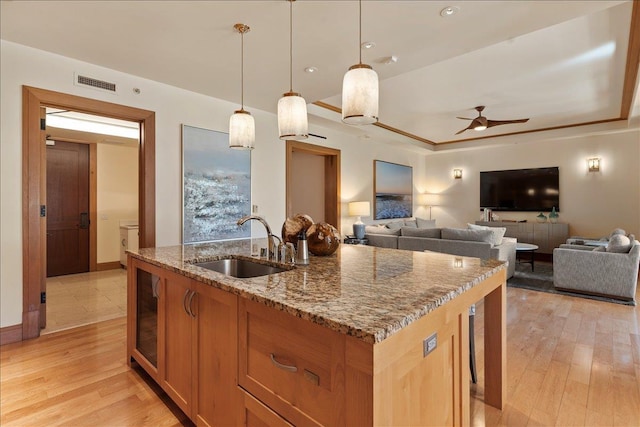 kitchen with light stone counters, ceiling fan, a kitchen island with sink, sink, and hanging light fixtures