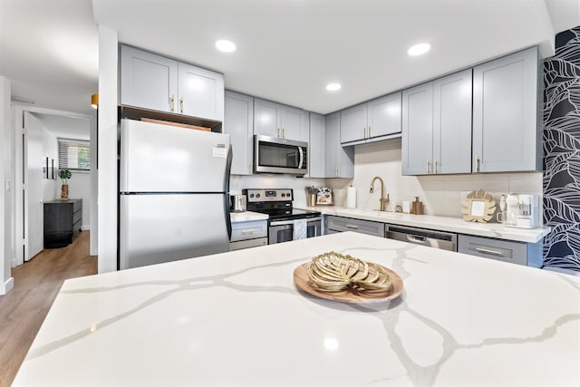 kitchen with light stone counters, appliances with stainless steel finishes, backsplash, light wood-type flooring, and gray cabinetry