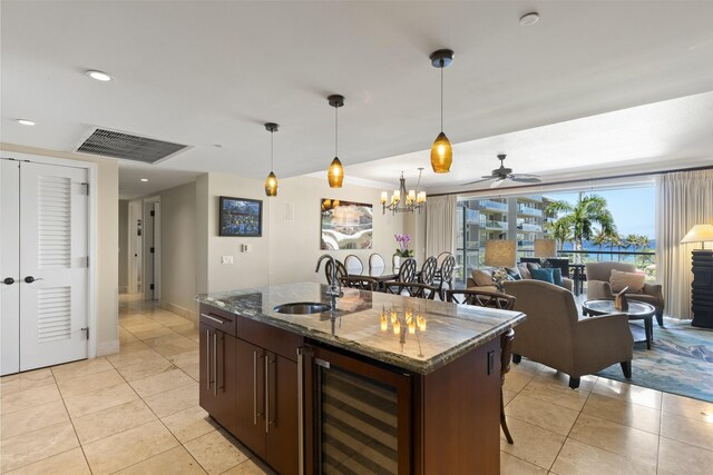 kitchen featuring ceiling fan with notable chandelier, beverage cooler, a kitchen island with sink, sink, and dark stone counters