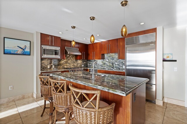 kitchen with built in appliances, light tile patterned floors, backsplash, an island with sink, and hanging light fixtures