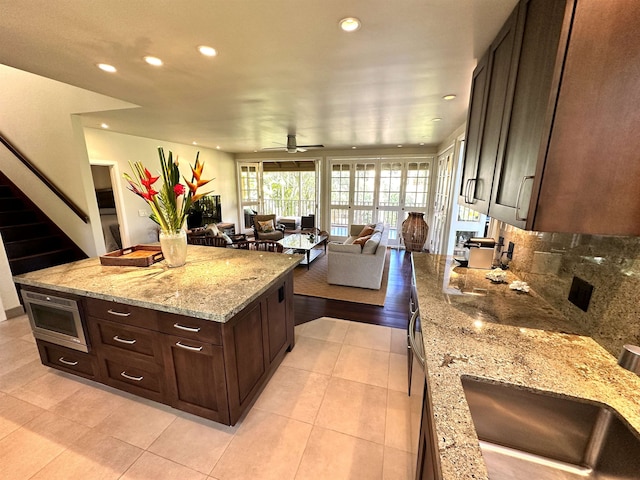 kitchen with ceiling fan, light tile floors, light stone counters, backsplash, and dark brown cabinetry