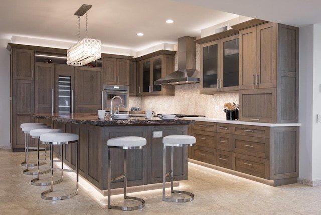 kitchen with pendant lighting, wall chimney exhaust hood, a kitchen bar, backsplash, and a center island with sink