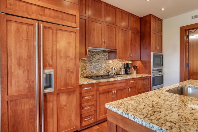 kitchen featuring under cabinet range hood, decorative backsplash, built in appliances, and brown cabinetry