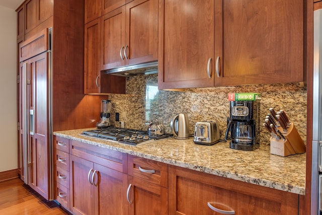 kitchen with backsplash, under cabinet range hood, light wood-type flooring, stainless steel gas stovetop, and paneled fridge