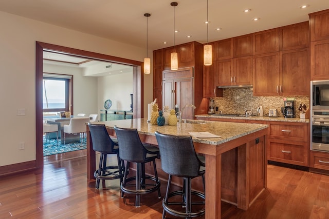 kitchen with wood finished floors, brown cabinets, a sink, built in appliances, and tasteful backsplash