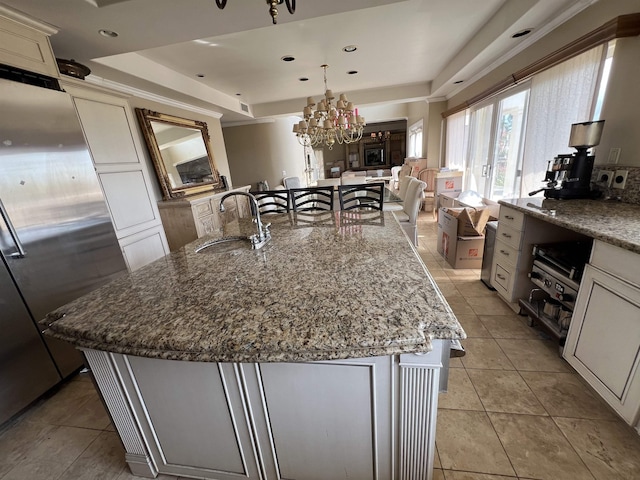 kitchen with an island with sink, a sink, refrigerator, a raised ceiling, and light stone countertops