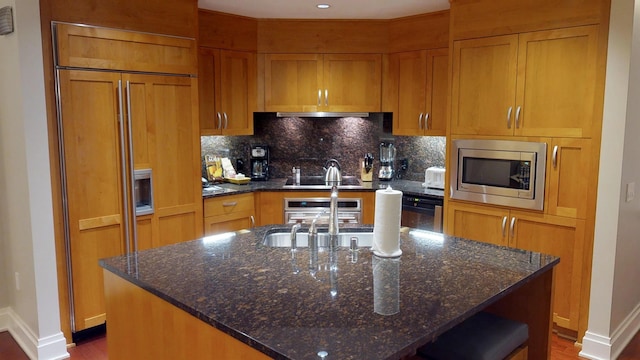 kitchen with dark stone countertops, backsplash, built in appliances, and a center island with sink