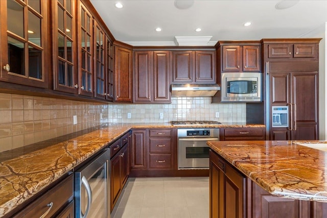 kitchen with tasteful backsplash, under cabinet range hood, built in appliances, beverage cooler, and stone counters