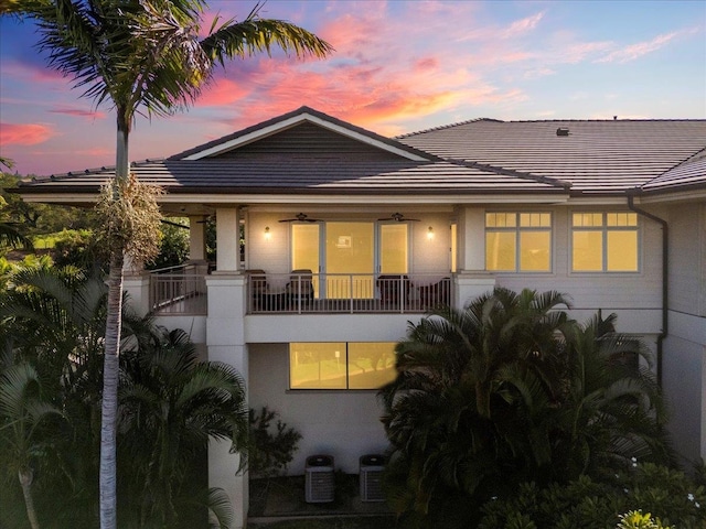 back of property at dusk featuring stucco siding, a balcony, central AC, and a tiled roof