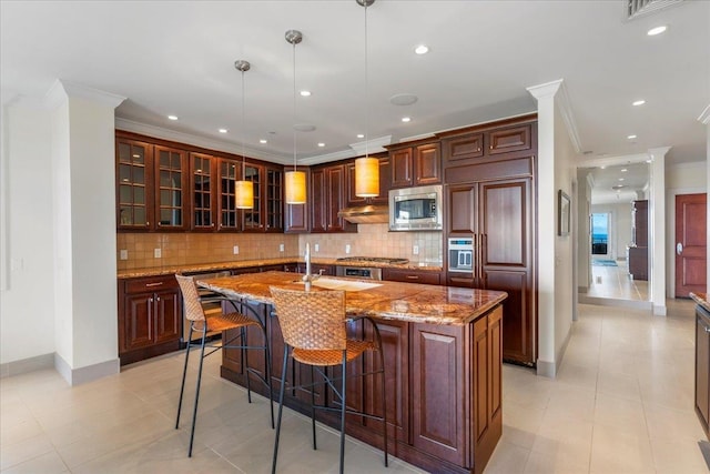 kitchen with a kitchen island with sink, ornamental molding, a kitchen breakfast bar, backsplash, and built in appliances