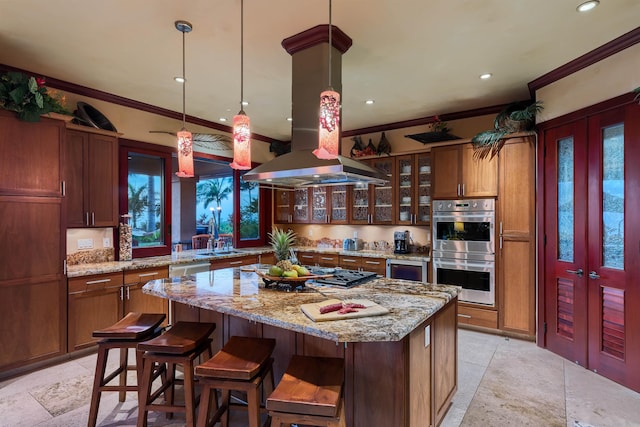 kitchen with hanging light fixtures, appliances with stainless steel finishes, a kitchen island, island exhaust hood, and light stone countertops