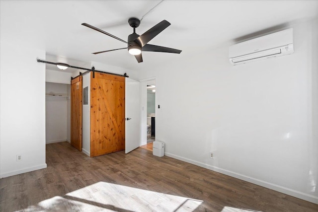 unfurnished bedroom with a walk in closet, a wall unit AC, a barn door, wood finished floors, and baseboards