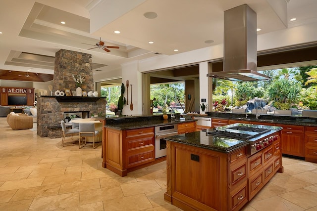 kitchen featuring ceiling fan, dark stone counters, island exhaust hood, a fireplace, and a center island