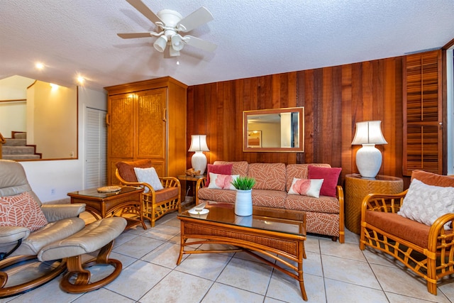 living room featuring a textured ceiling, ceiling fan, light tile patterned floors, wood walls, and stairway