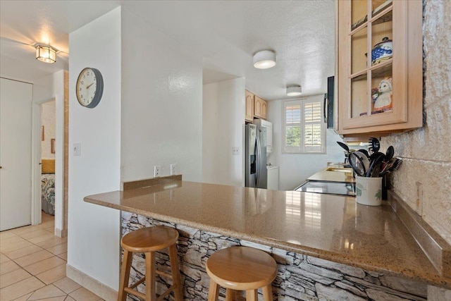 kitchen with kitchen peninsula, light brown cabinetry, stainless steel fridge with ice dispenser, light tile patterned floors, and a kitchen breakfast bar