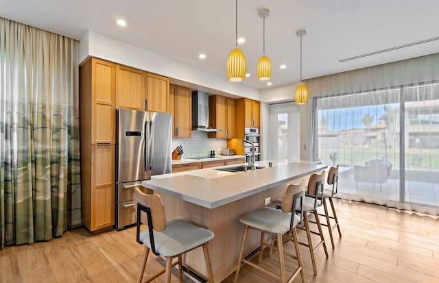 kitchen with wall chimney exhaust hood, hanging light fixtures, a kitchen breakfast bar, stainless steel appliances, and light hardwood / wood-style floors