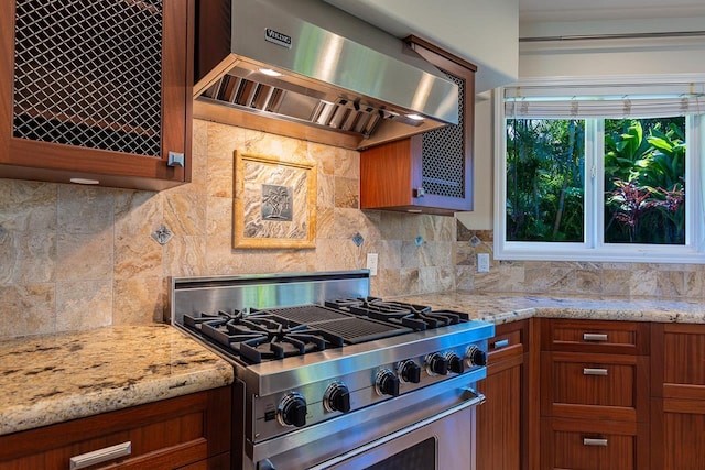 kitchen featuring brown cabinetry, light stone counters, gas stove, backsplash, and range hood