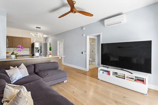 living area featuring ceiling fan with notable chandelier, baseboards, light wood-style floors, and a wall unit AC