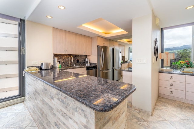 kitchen with appliances with stainless steel finishes, backsplash, dark stone countertops, light brown cabinets, and kitchen peninsula