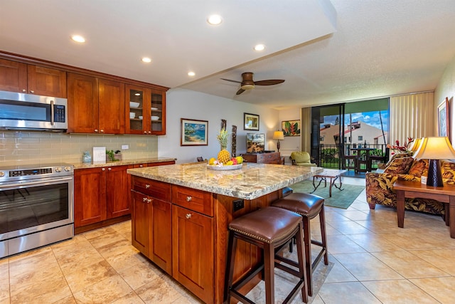kitchen with glass insert cabinets, a kitchen bar, light stone countertops, backsplash, and appliances with stainless steel finishes