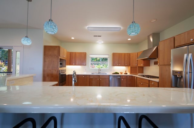 kitchen with wall chimney exhaust hood, light stone counters, sink, hanging light fixtures, and appliances with stainless steel finishes