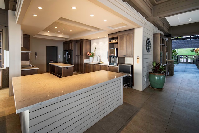 kitchen with light stone counters, sink, a large island, black refrigerator with ice dispenser, and a tray ceiling