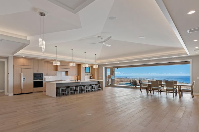 kitchen with a raised ceiling, black appliances, a water view, a center island with sink, and hanging light fixtures