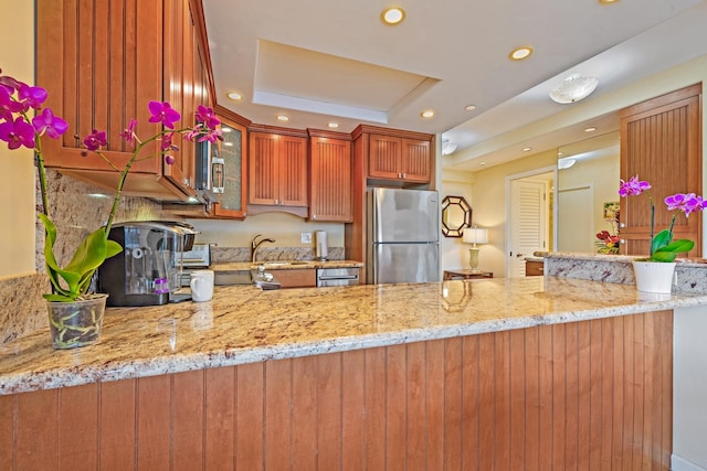 kitchen with stainless steel refrigerator, kitchen peninsula, a raised ceiling, light stone countertops, and sink