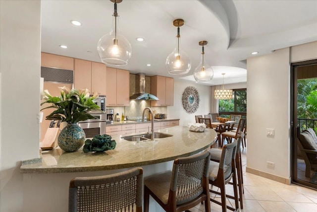 kitchen with light brown cabinets, hanging light fixtures, sink, kitchen peninsula, and wall chimney exhaust hood