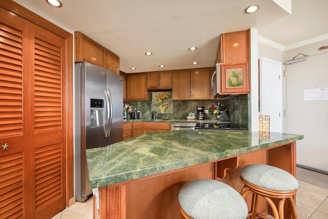 kitchen with light tile patterned floors, a peninsula, decorative backsplash, brown cabinetry, and stainless steel fridge