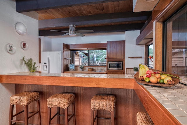 kitchen featuring white fridge with ice dispenser, tile counters, stainless steel microwave, beam ceiling, and ceiling fan