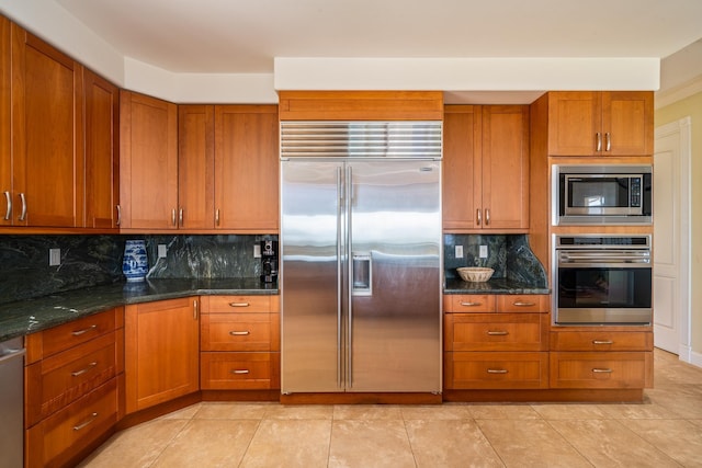 kitchen with light tile patterned floors, built in appliances, backsplash, and dark stone countertops