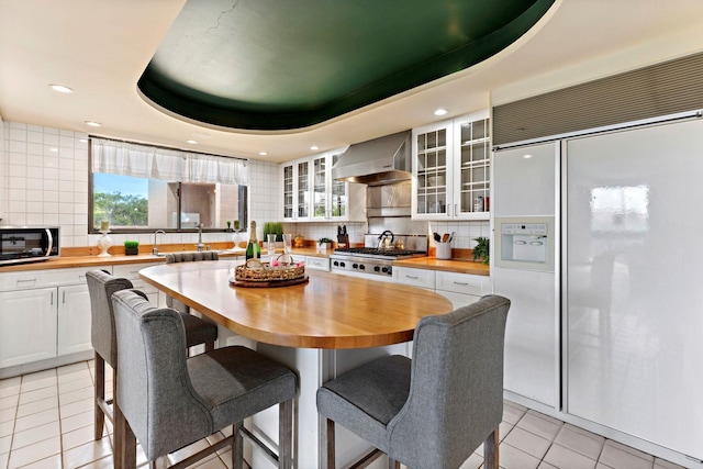 kitchen featuring wall chimney exhaust hood, stainless steel gas cooktop, a raised ceiling, built in fridge, and white cabinets