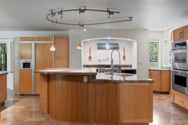 kitchen with wall chimney exhaust hood, paneled built in refrigerator, pendant lighting, stainless steel double oven, and a kitchen island with sink