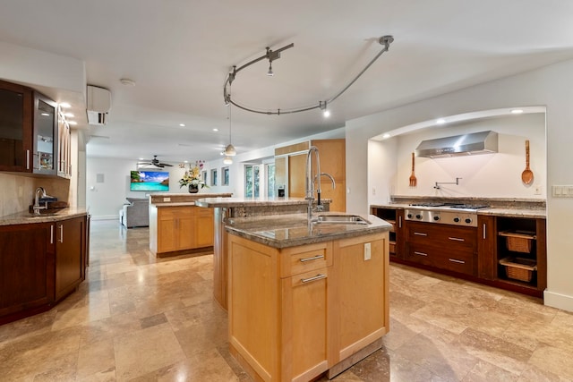 kitchen with light stone counters, sink, a center island with sink, wall chimney range hood, and stainless steel gas stovetop