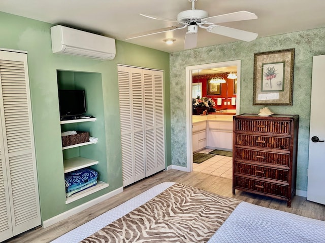 bedroom with a wall unit AC, connected bathroom, ceiling fan, and light hardwood / wood-style flooring