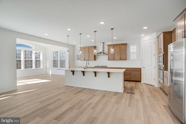 kitchen with a breakfast bar area, light countertops, appliances with stainless steel finishes, wall chimney range hood, and an island with sink