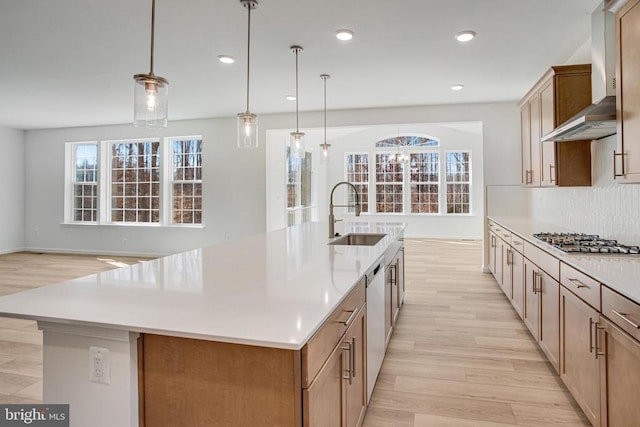 kitchen with a sink, light countertops, appliances with stainless steel finishes, wall chimney exhaust hood, and a large island with sink