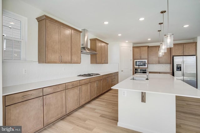 kitchen featuring a sink, light countertops, appliances with stainless steel finishes, wall chimney exhaust hood, and a center island with sink