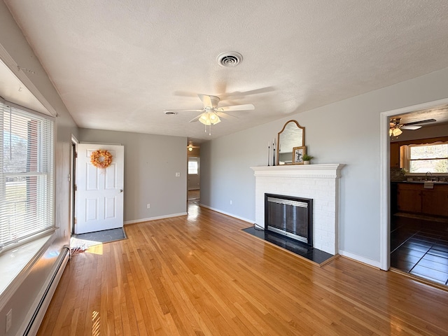 unfurnished living room with visible vents, light wood-type flooring, a fireplace, a ceiling fan, and a baseboard radiator