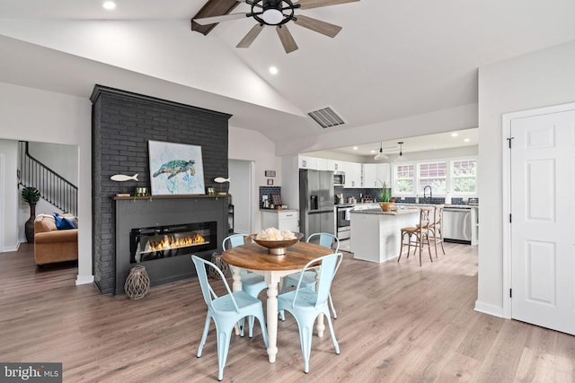 dining area with a ceiling fan, visible vents, light wood finished floors, a glass covered fireplace, and vaulted ceiling with beams