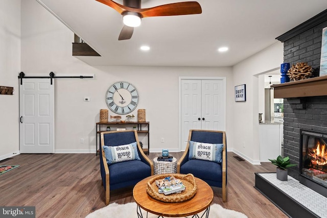living area featuring ceiling fan, a brick fireplace, a barn door, wood finished floors, and baseboards