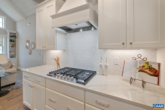 kitchen featuring custom exhaust hood, tasteful backsplash, stainless steel gas stovetop, lofted ceiling with beams, and light wood-type flooring