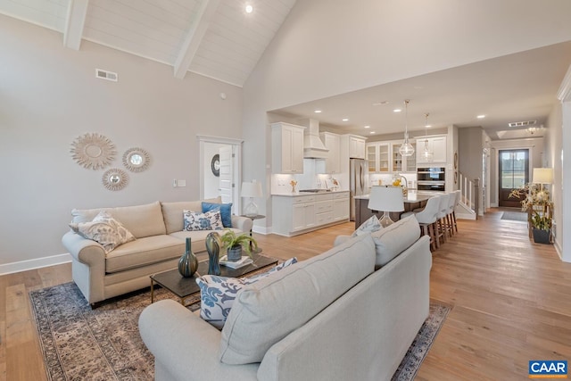 living room featuring light wood finished floors, visible vents, high vaulted ceiling, beamed ceiling, and baseboards