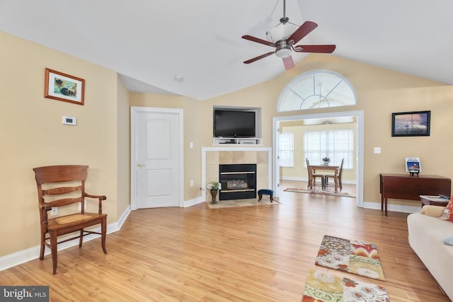 living area featuring baseboards, lofted ceiling, light wood-style flooring, a tile fireplace, and a ceiling fan