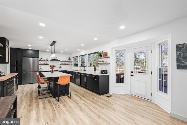 kitchen featuring a sink, a kitchen island, dark cabinetry, stainless steel refrigerator, and open shelves