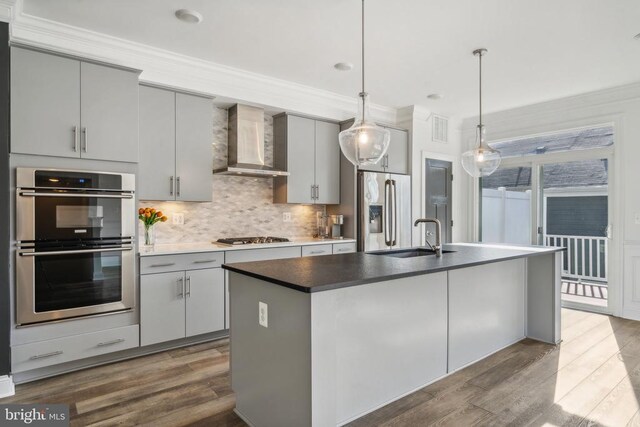 kitchen featuring sink, hanging light fixtures, stainless steel appliances, a kitchen island with sink, and wall chimney range hood